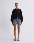 A model stands against a light gray studio background wearing a black zip-up bomber-style sweatshirt, blue denim shorts, and black sandals.