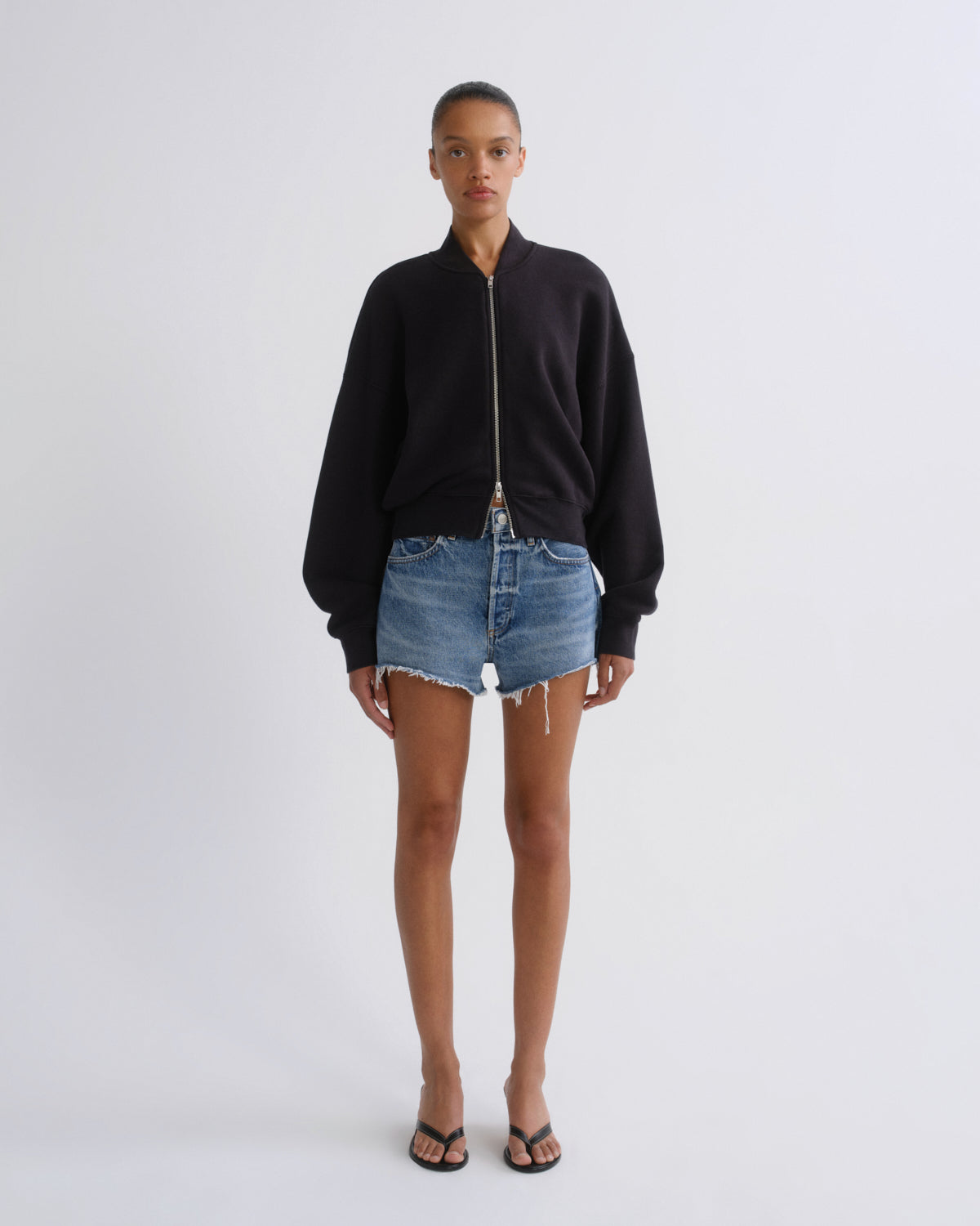 A model stands against a light gray studio background wearing a black zip-up bomber-style sweatshirt, blue denim shorts, and black sandals.