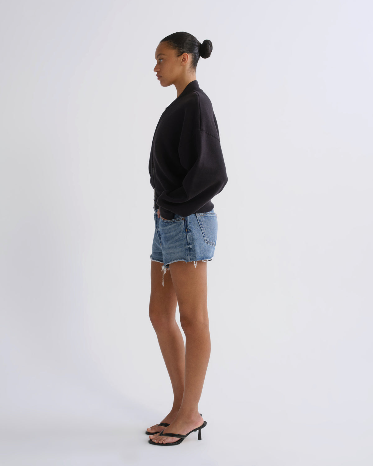 A woman in profile wearing a black crewneck sweatshirt, light-wash denim shorts, and black heeled sandals, standing against a plain light gray studio background.