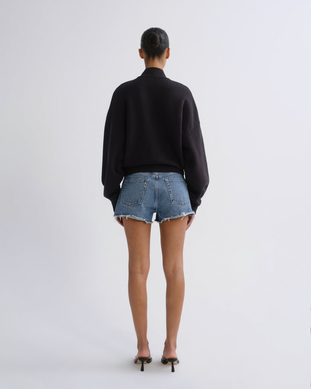 A model viewed from behind wearing a black long-sleeve mock-neck sweater, light-wash denim frayed-hem shorts, and black high-heeled sandals against a plain light gray studio background.