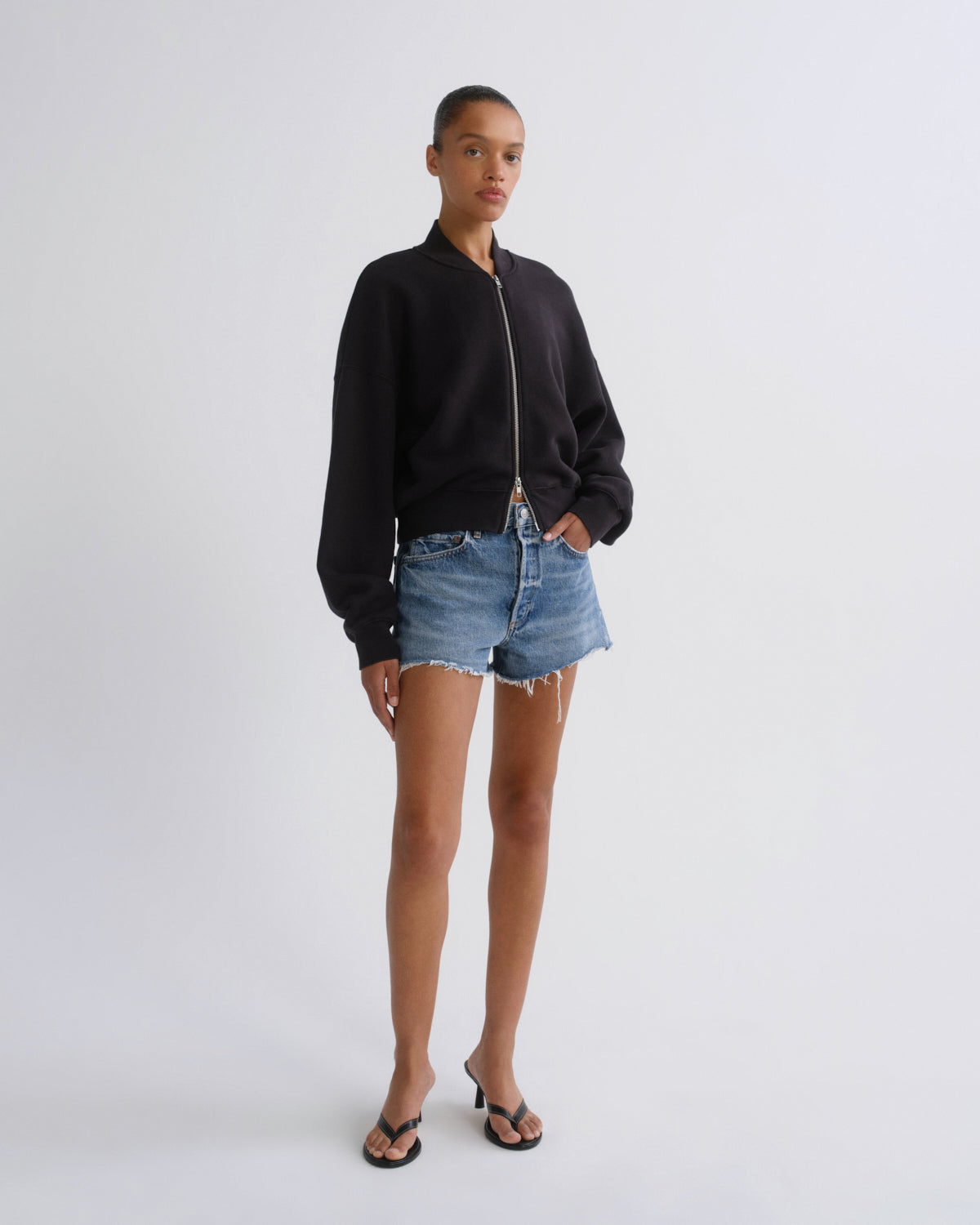 A model stands against a light gray studio background wearing a black zip-up bomber jacket, light-wash denim shorts, and black flip-flop sandals.