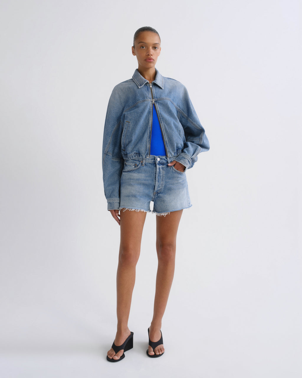 A model stands against a light gray studio background wearing a light-wash blue denim jacket and matching frayed denim shorts over a bright blue top, with black sandals.