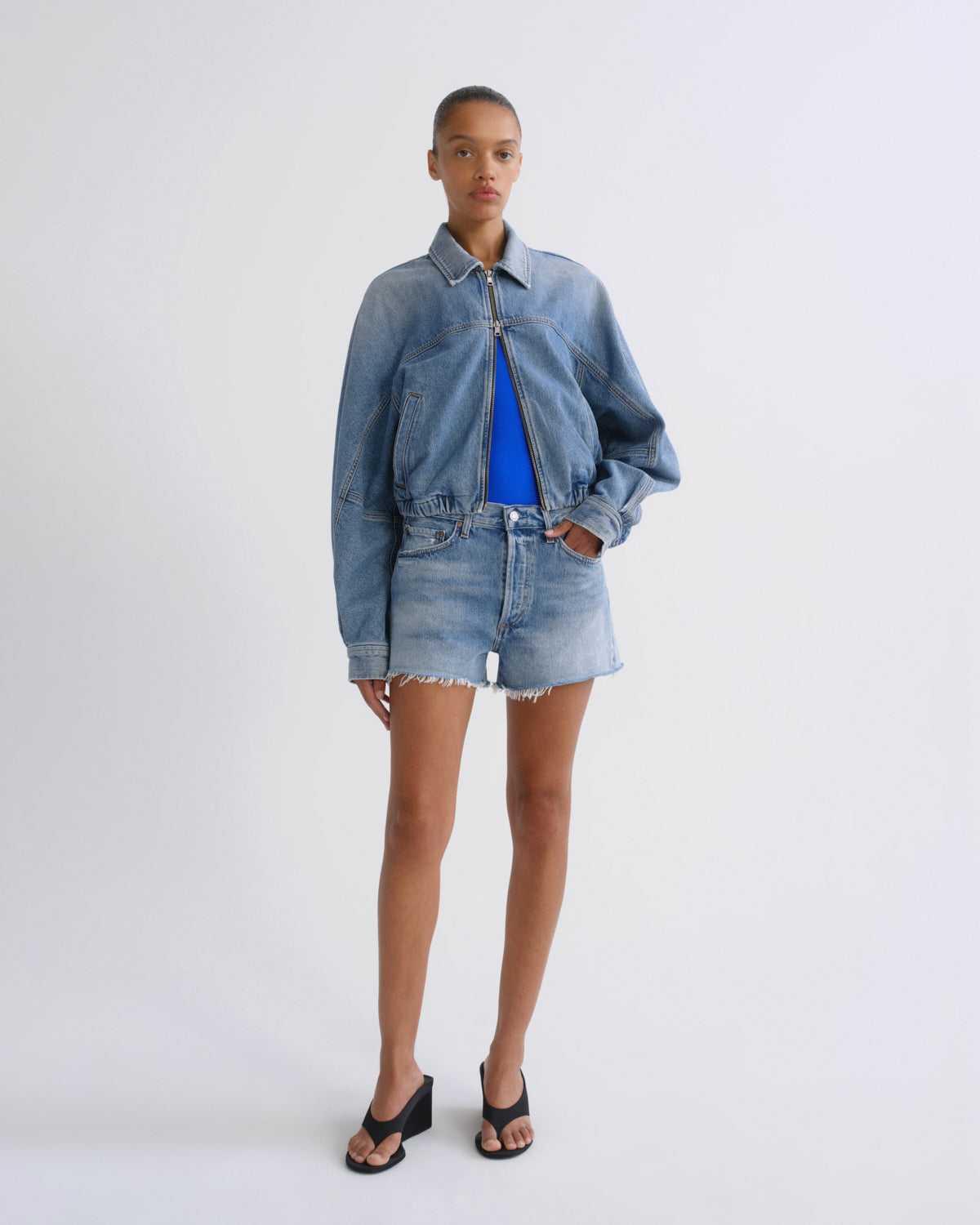 A model stands against a light gray studio background wearing a light-wash blue denim jacket and matching frayed denim shorts over a bright blue top, with black sandals.