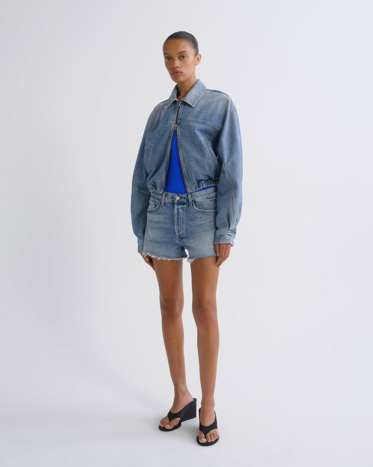 A model stands against a light gray studio background wearing a light-wash blue denim jacket and matching denim shorts over a bright blue top, with black slide sandals.