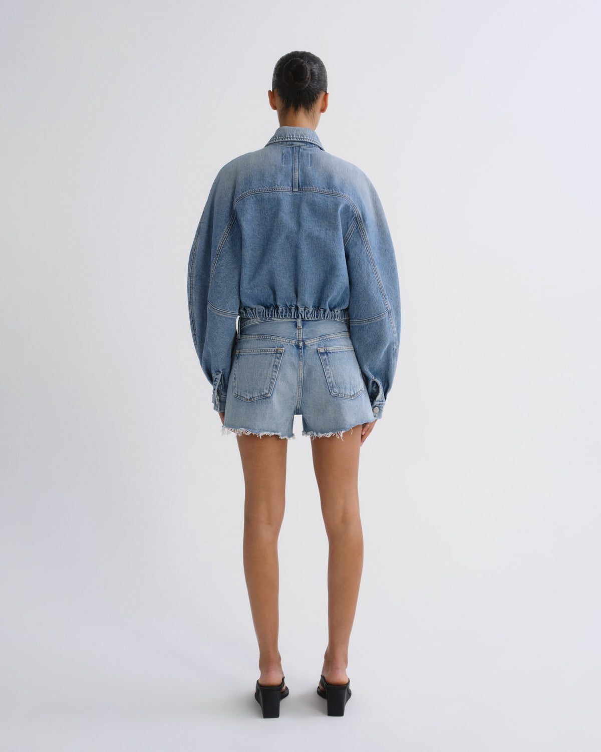 A model viewed from the back wearing a light blue denim cropped jacket and matching frayed-hem denim shorts, paired with black mule heels, against a plain white studio background.