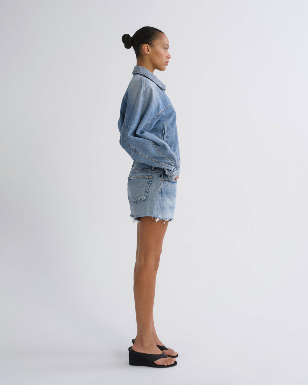 A model standing in profile wearing a light blue denim jacket and matching denim mini skirt, paired with black wedge sandals, against a plain light gray studio background.