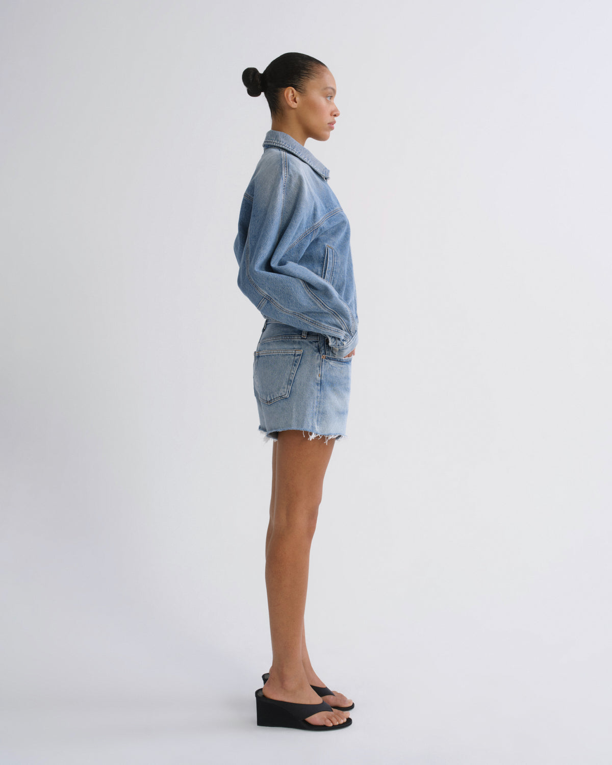 A model standing in profile wearing a light blue denim jacket and matching denim mini skirt, paired with black wedge sandals, against a plain light gray studio background.