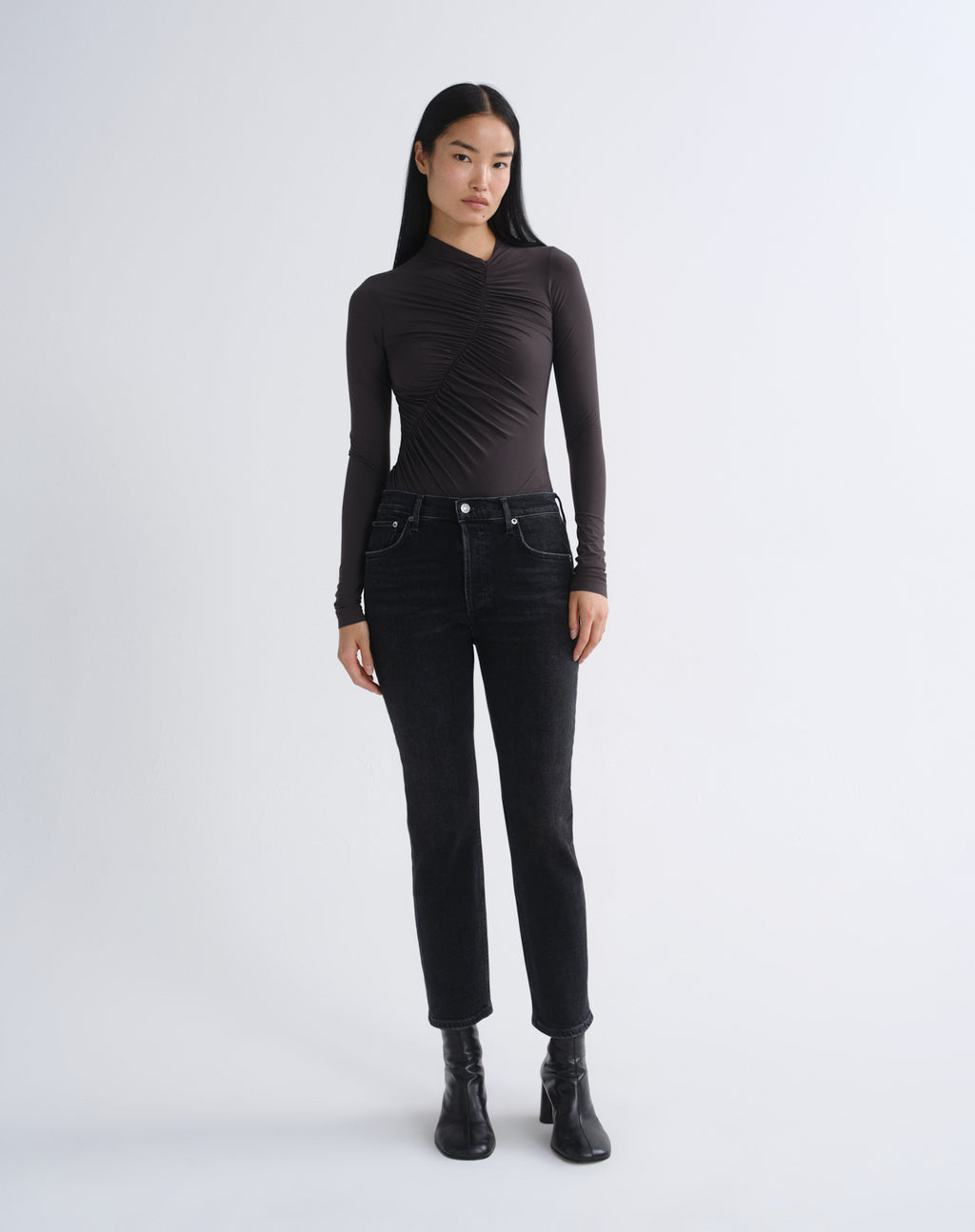 A model stands against a light gray studio background wearing a fitted black long-sleeve top with ruched detailing, black jeans, and black ankle boots.