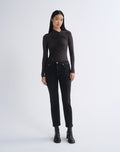 A model stands against a light gray studio background wearing a fitted black long-sleeve top with ruched detailing, black jeans, and black ankle boots.