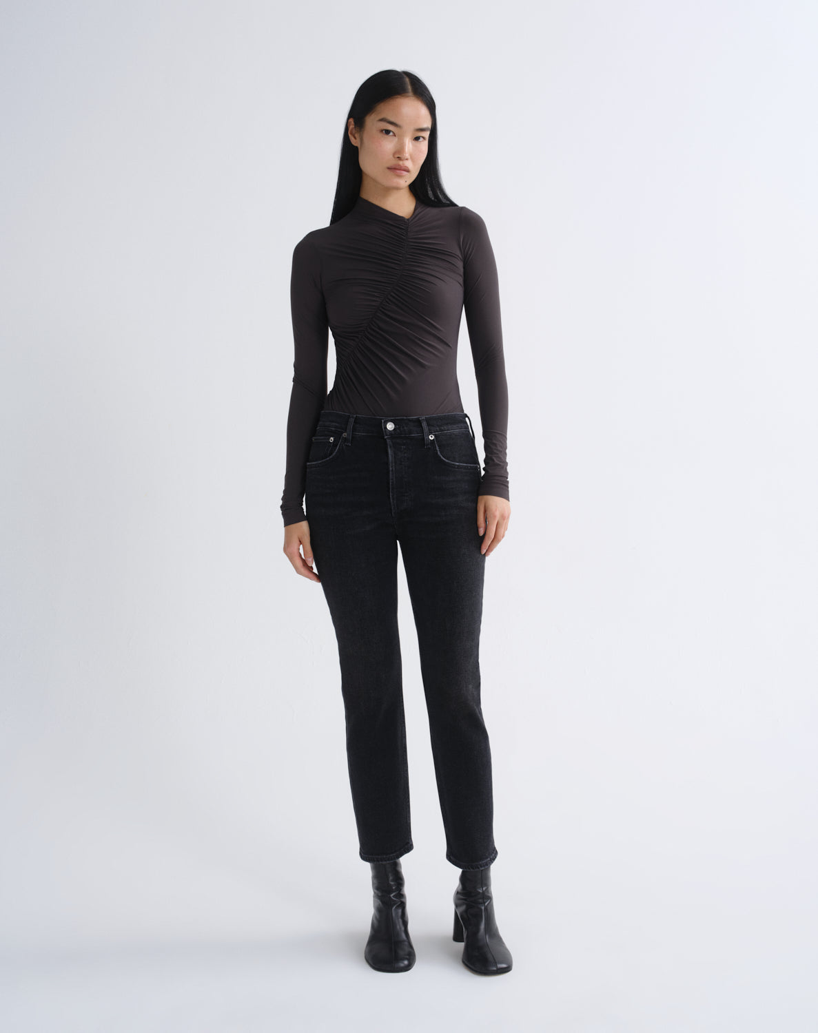 A model stands against a light gray studio background wearing a fitted black long-sleeve top with ruched detailing, black jeans, and black ankle boots.