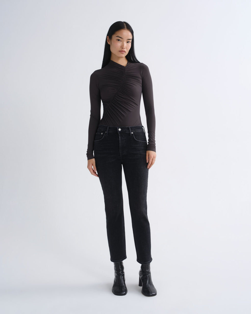 A model stands against a light gray studio background wearing a fitted black long-sleeve top with ruched detailing, black jeans, and black ankle boots.