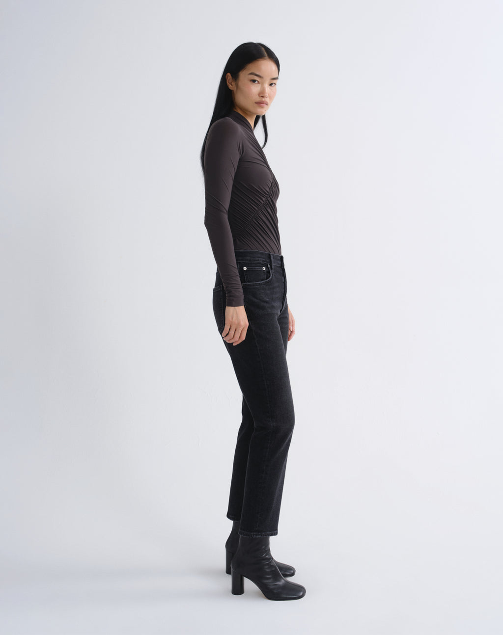 A model is shown in profile wearing a dark brown ribbed long-sleeve top, black cropped jeans, and black heeled ankle boots against a light gray studio background.