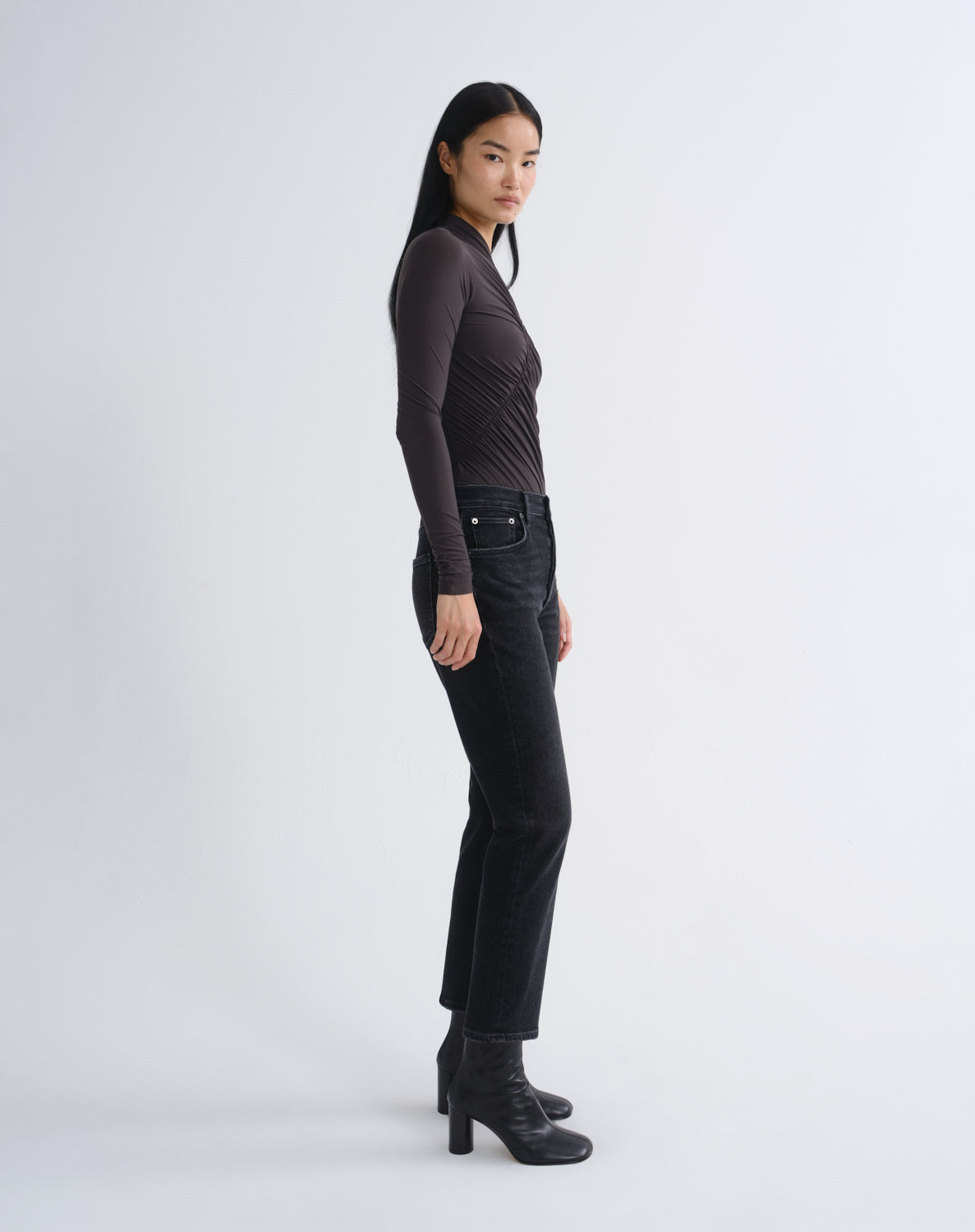 A model is shown in profile wearing a dark brown ribbed long-sleeve top, black cropped jeans, and black heeled ankle boots against a light gray studio background.