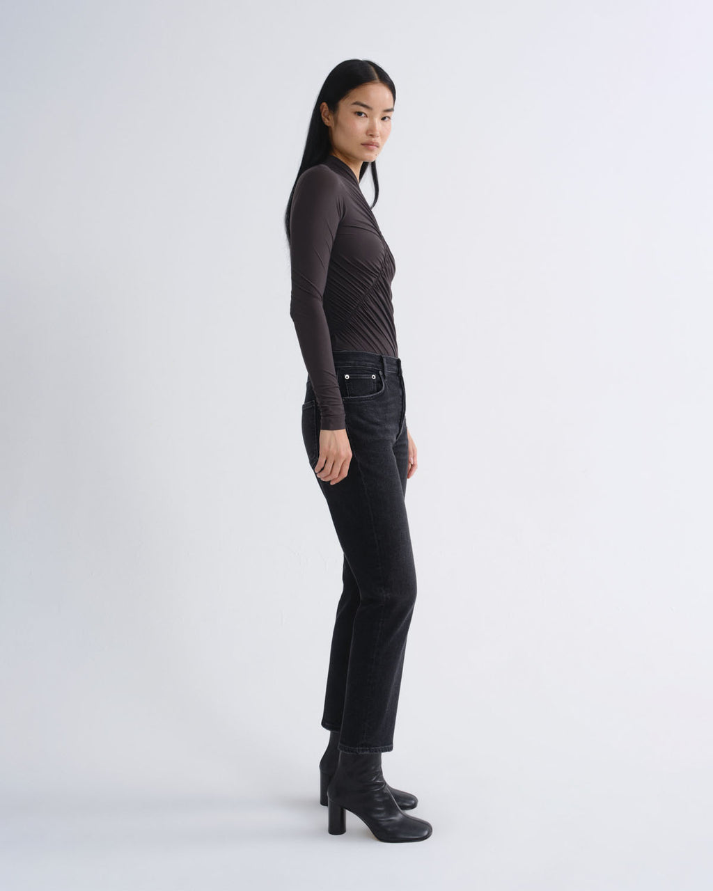 A model is shown in profile wearing a dark brown ribbed long-sleeve top, black high-waisted straight-leg jeans, and black leather ankle boots against a light gray studio background.