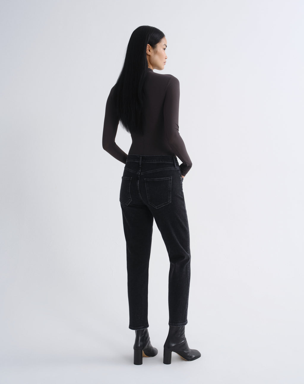A woman with long black hair is shown from the back wearing a dark brown long-sleeve top, black high-waisted jeans, and black ankle boots, standing against a light gray studio background.