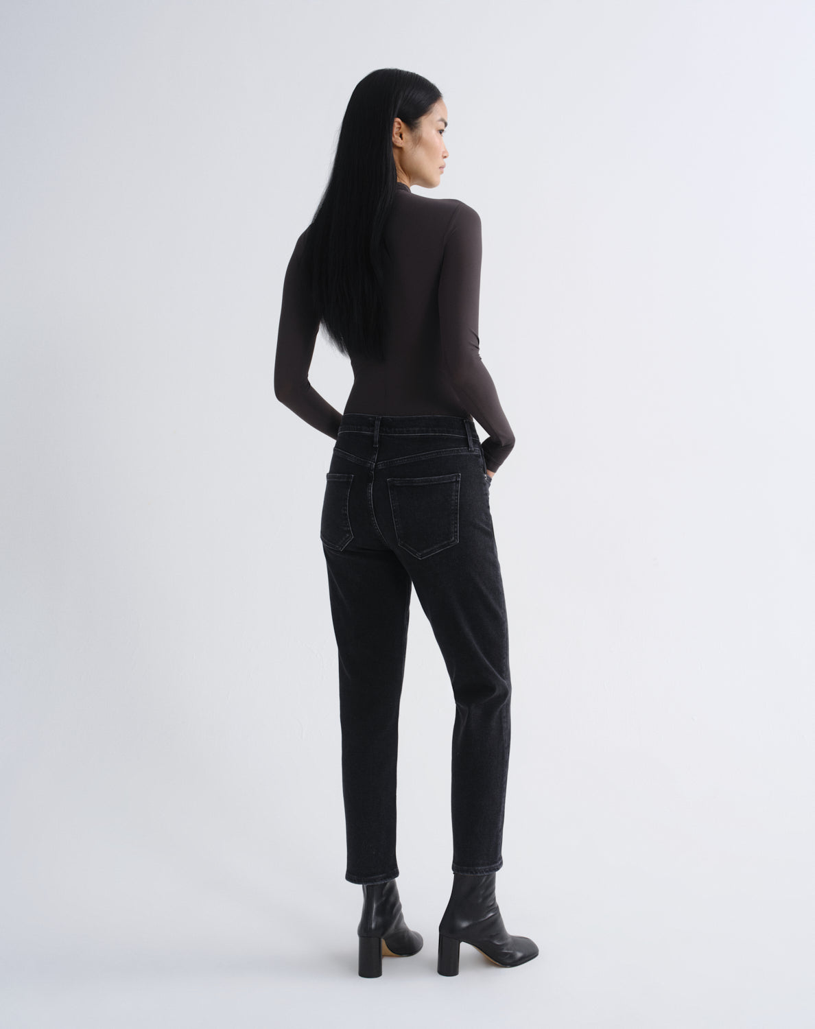 A woman with long black hair is shown from the back wearing a dark brown long-sleeve top, black high-waisted jeans, and black ankle boots, standing against a light gray studio background.
