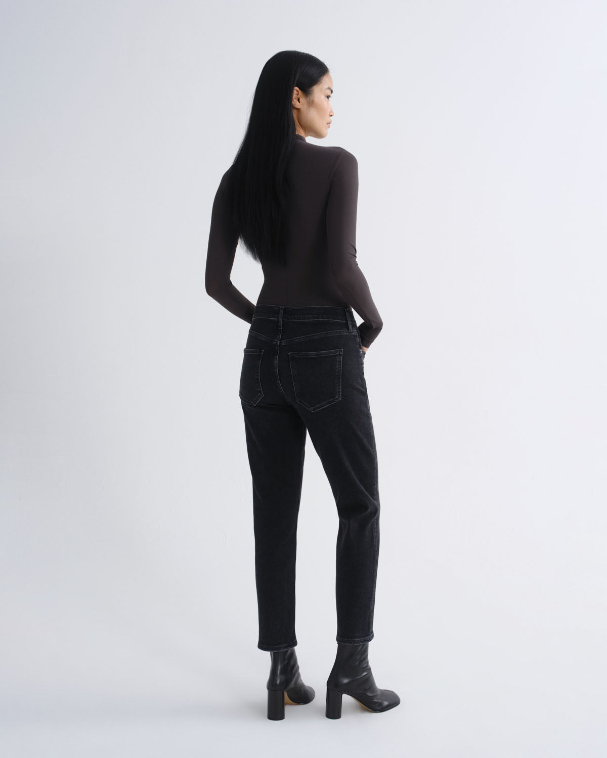 A woman with long black hair is shown from the back wearing a dark brown long-sleeve top, black high-waisted jeans, and black ankle boots, standing against a light gray studio background.