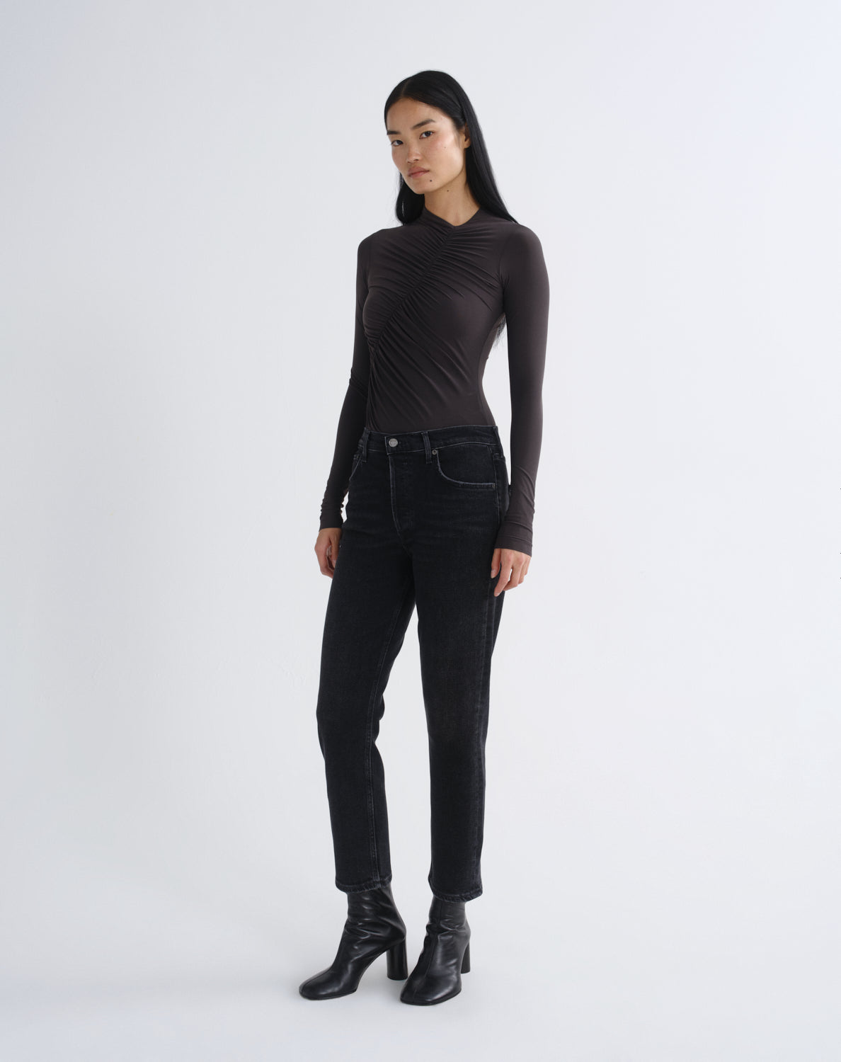 A woman modeling a dark brown long-sleeve top with a unique draped, gathered neckline, paired with black jeans and black leather ankle boots, against a light gray studio background.