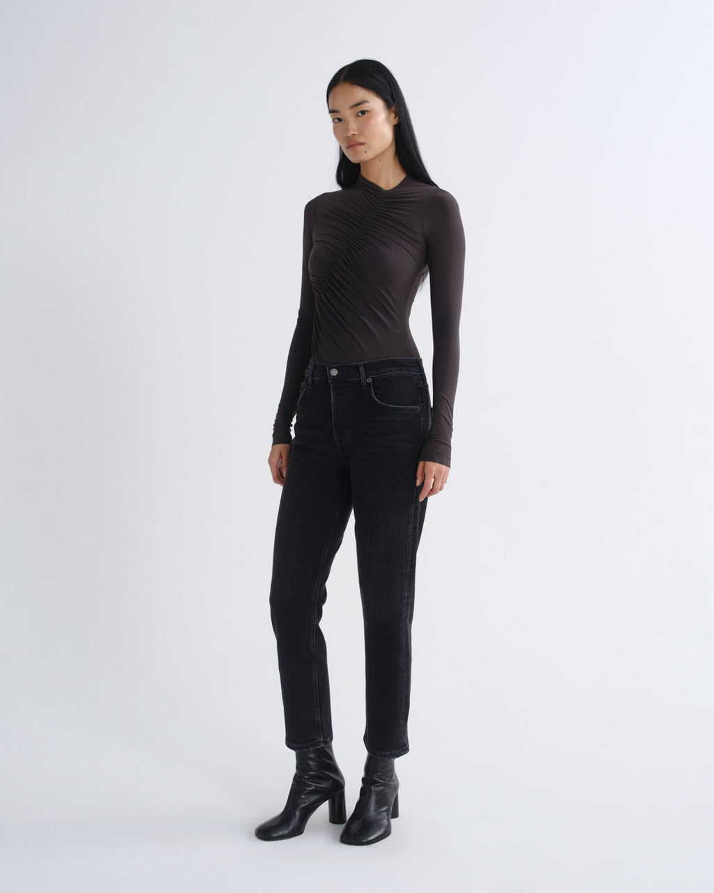 A model stands against a light gray studio background wearing a dark brown long-sleeve ribbed mock-neck top, black jeans, and black heeled ankle boots.