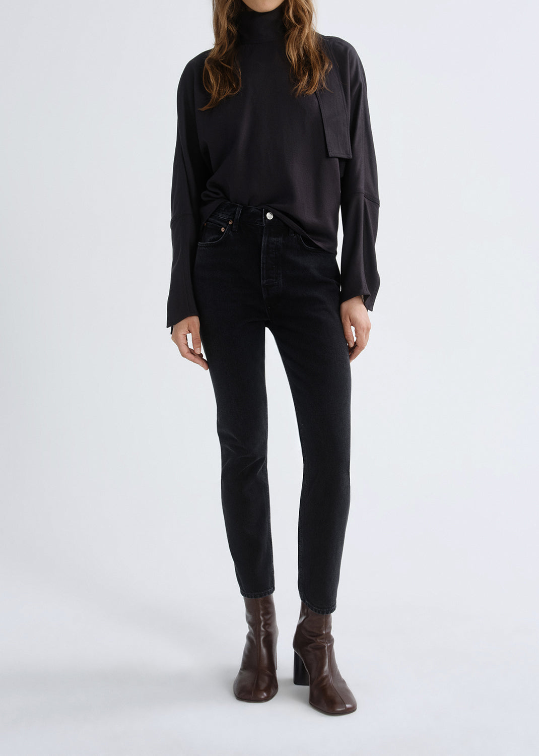 Person wearing a black long sleeve turtleneck sweater with black skinny jeans and brown ankle boots on a grey background