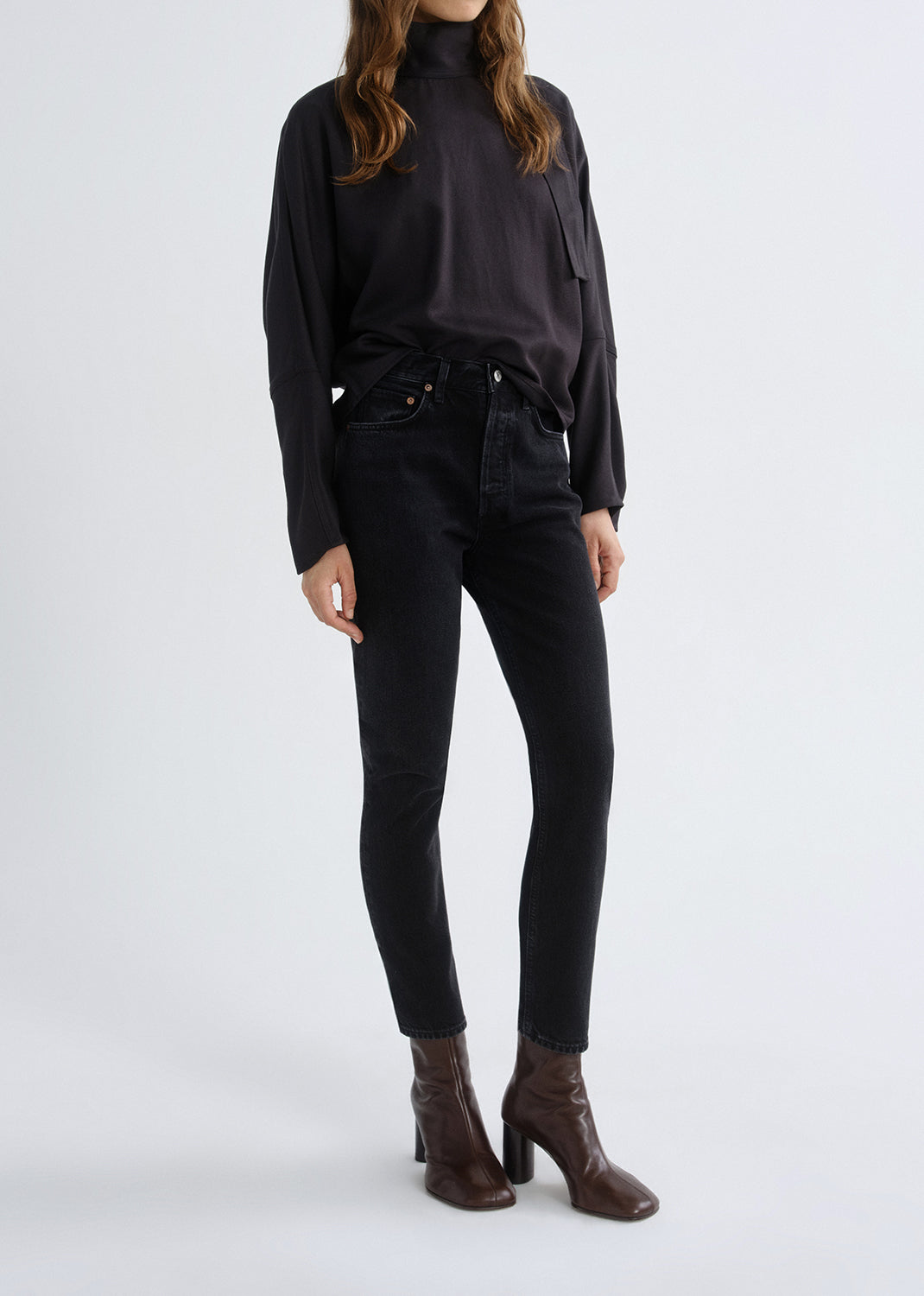 Person wearing a black long sleeve turtleneck sweater with black skinny jeans and brown ankle boots on a grey background