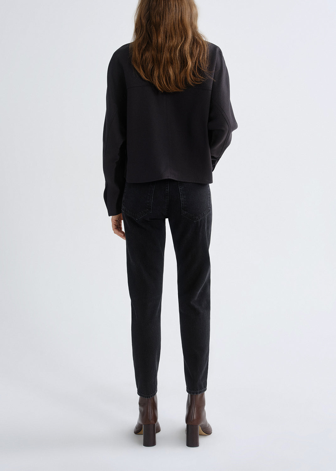 Person wearing a black long sleeve turtleneck sweater with black skinny jeans viewed from the back on a grey background