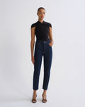 A model stands against a light gray studio background wearing a black short-sleeve top with a gathered neckline, dark blue cropped jeans, and black strappy heels.