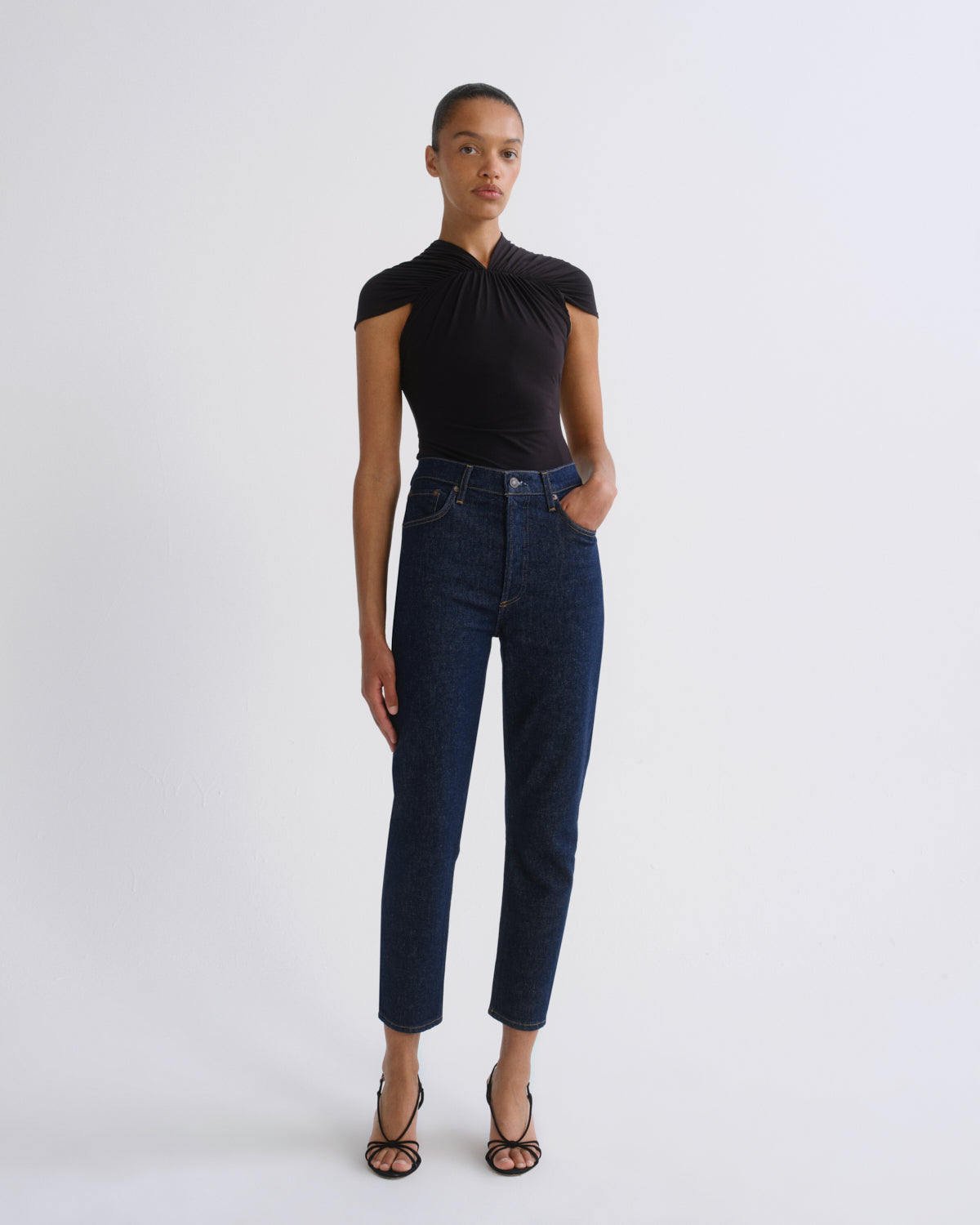 A model stands against a light gray studio background wearing a black short-sleeve top with a gathered neckline, dark blue cropped jeans, and black strappy heels.