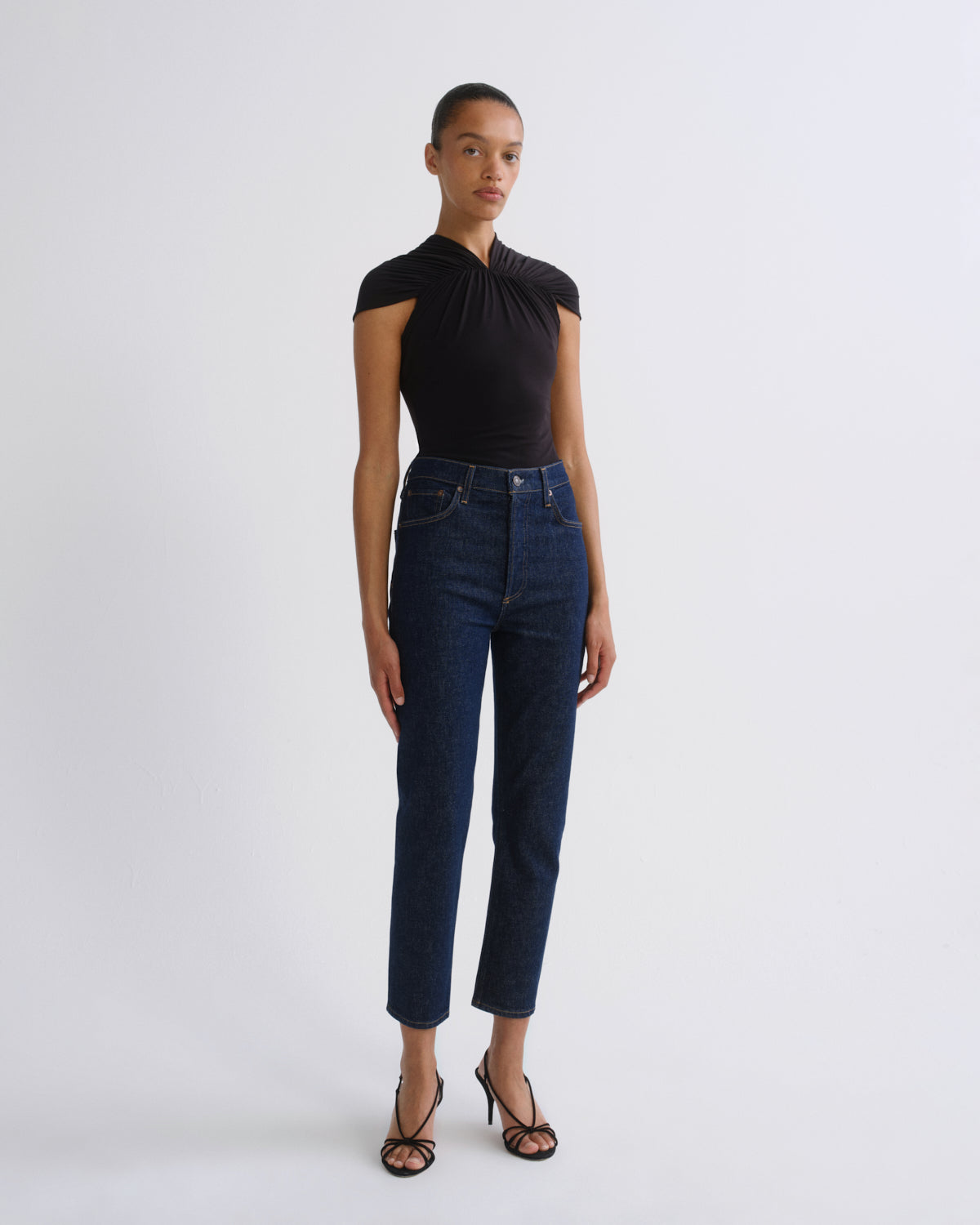 A model stands against a light gray studio background wearing a black sleeveless top with a draped neckline and dark blue cropped jeans with black strappy heels.