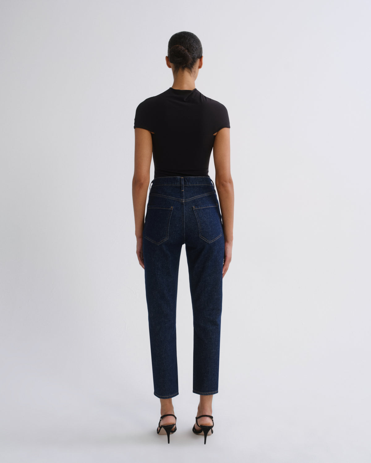 A model shown from the back wearing a black short-sleeve mock neck top, dark blue high-waisted straight-leg jeans, and black strappy heels against a light gray studio background.