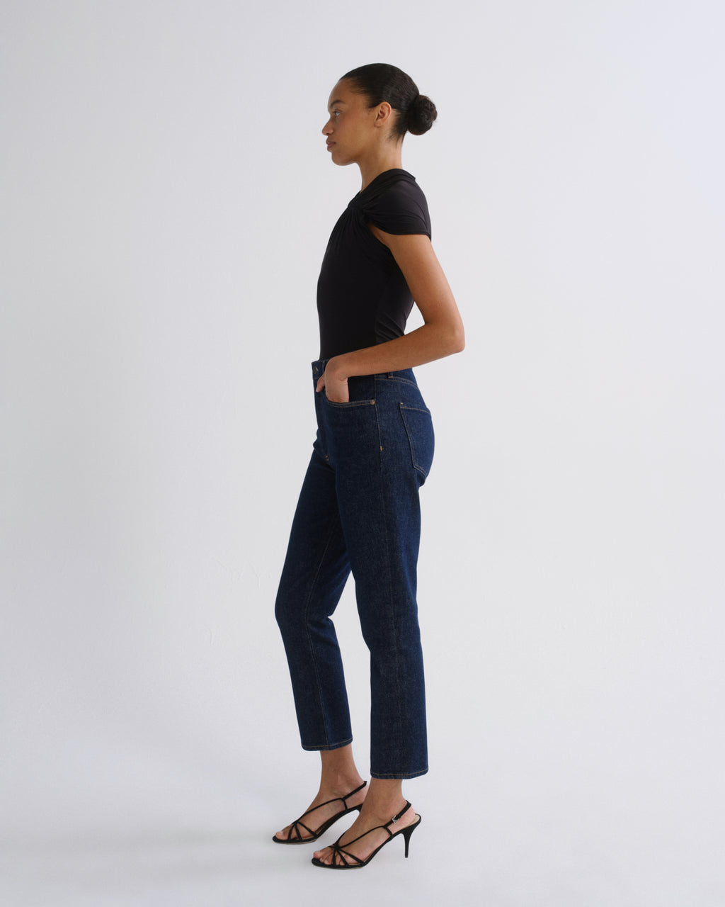 A woman in a black short-sleeve top and dark blue cropped jeans stands in profile against a light gray studio background, wearing black strappy high-heeled sandals.