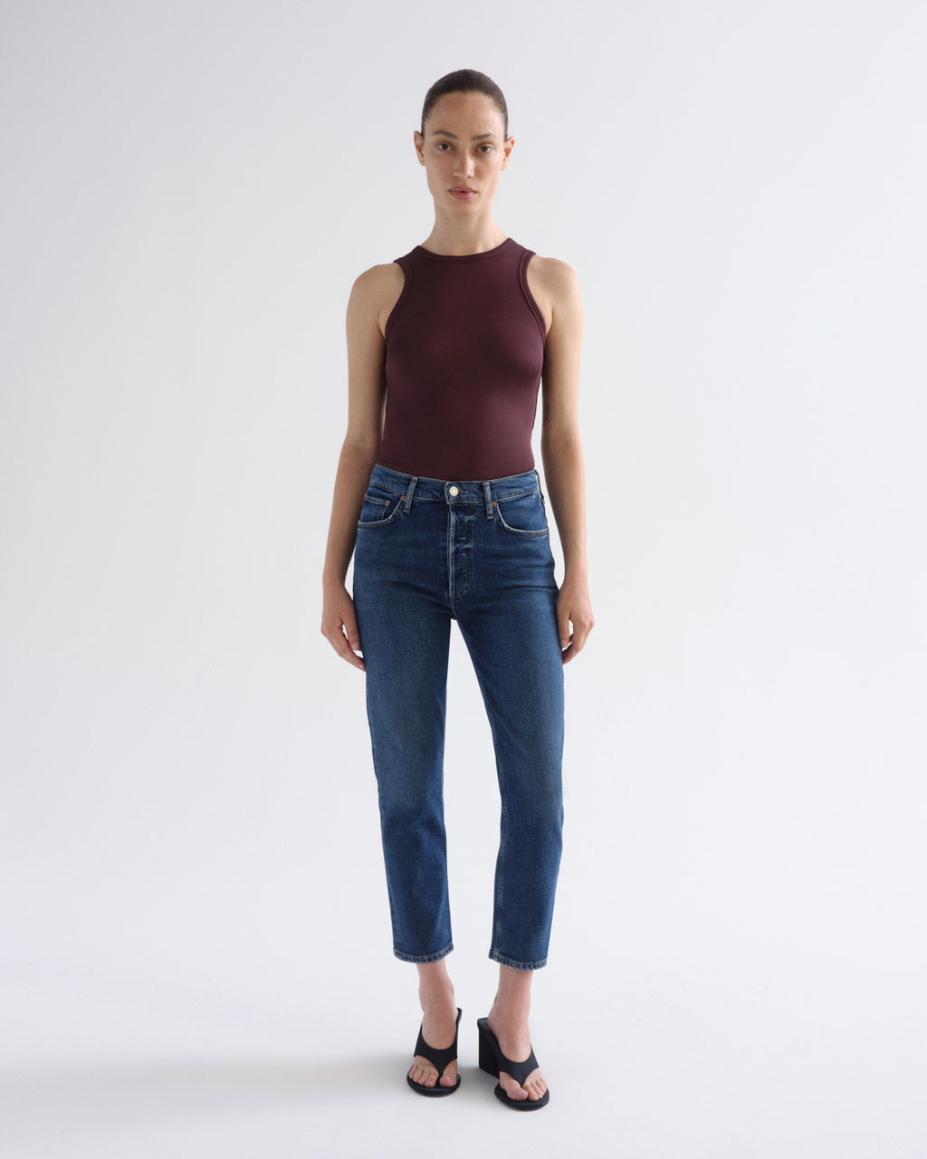 A model stands against a light gray studio background wearing a sleeveless dark burgundy top, blue cropped jeans, and black sandals.