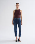 A model stands against a light gray studio background wearing a sleeveless dark burgundy top, blue cropped jeans, and black sandals.
