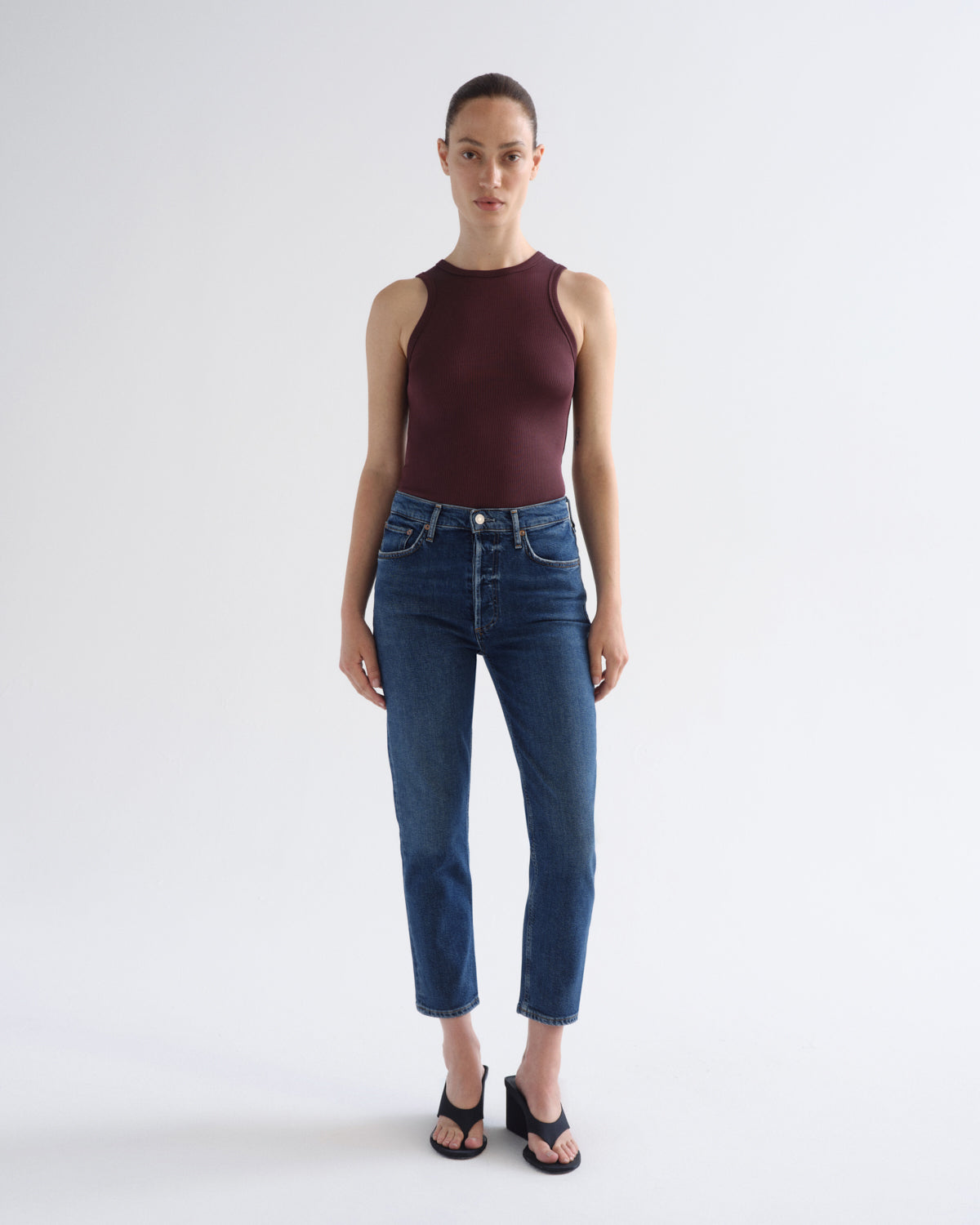 A model stands against a light gray studio background wearing a sleeveless dark burgundy top, blue cropped jeans, and black sandals.