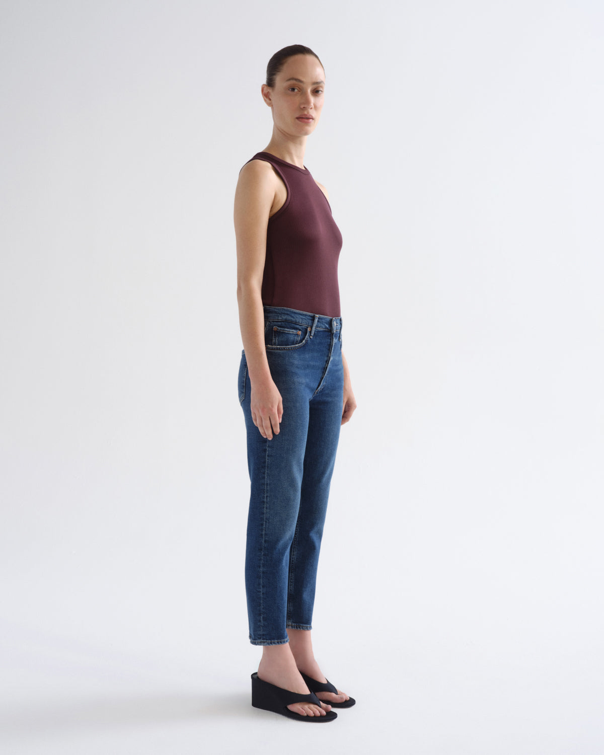 A woman stands in profile wearing a sleeveless dark burgundy top, blue cropped jeans, and black wedge sandals against a clean light gray studio background.