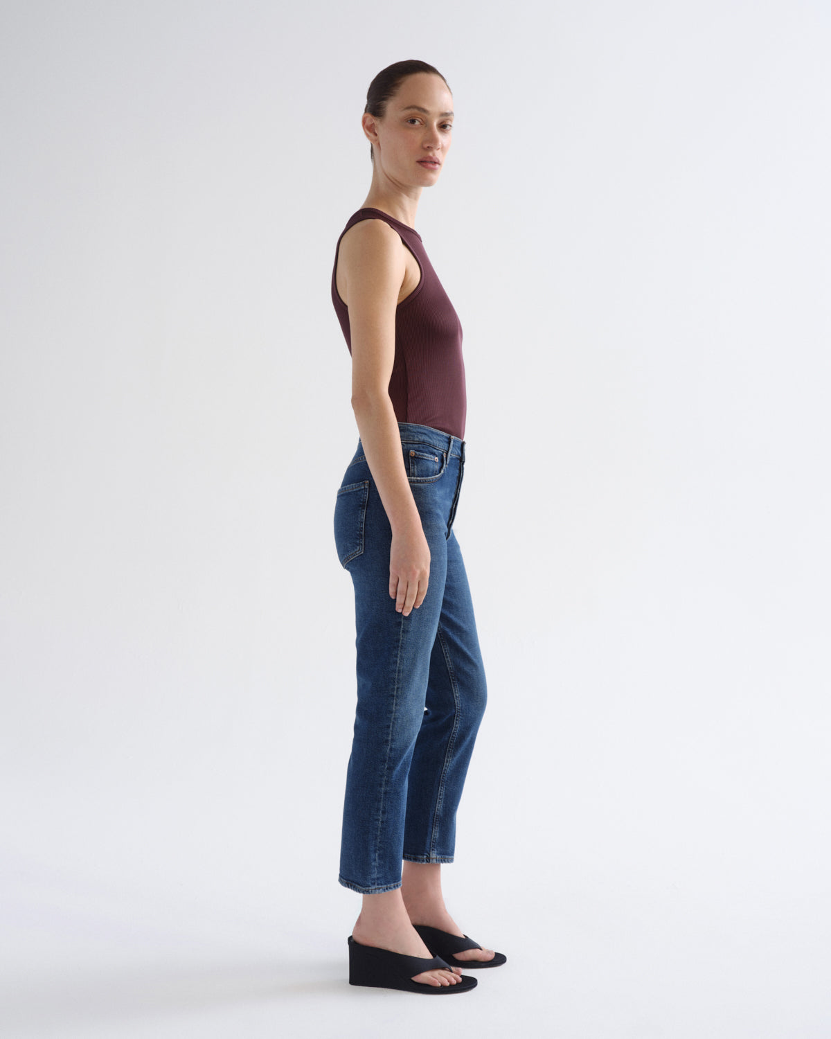 A woman stands in profile wearing a sleeveless dark burgundy top, dark blue cropped jeans, and black wedge sandals against a clean white studio background.