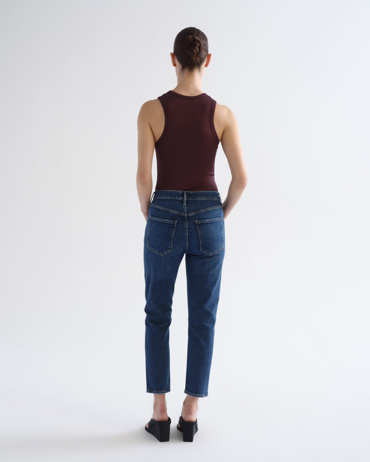 A model shown from the back wearing a sleeveless dark burgundy top, blue cropped jeans, and black heels against a plain white studio background.