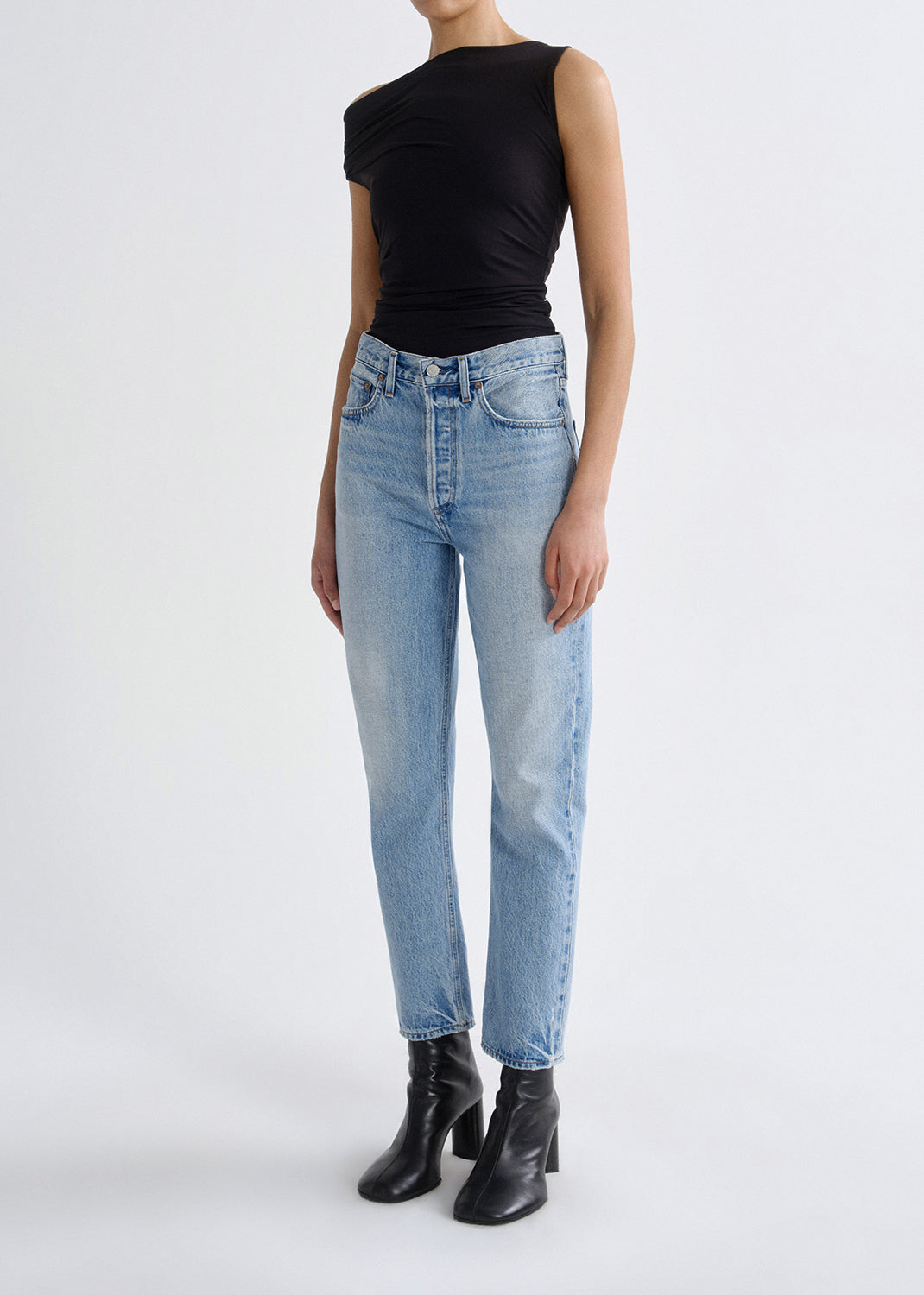 A model wearing light-wash blue high-rise jeans, a black sleeveless top, and black leather ankle boots, photographed against a seamless light gray studio background.