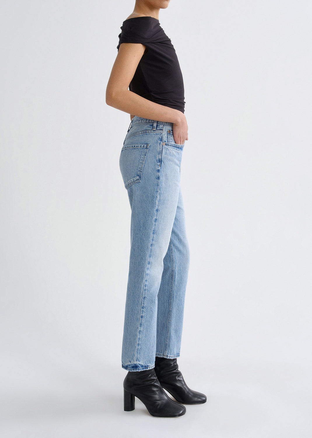 A side-profile view of a model wearing light-wash blue straight-leg jeans, a black off-the-shoulder top, and black heeled ankle boots against a plain light gray studio background.