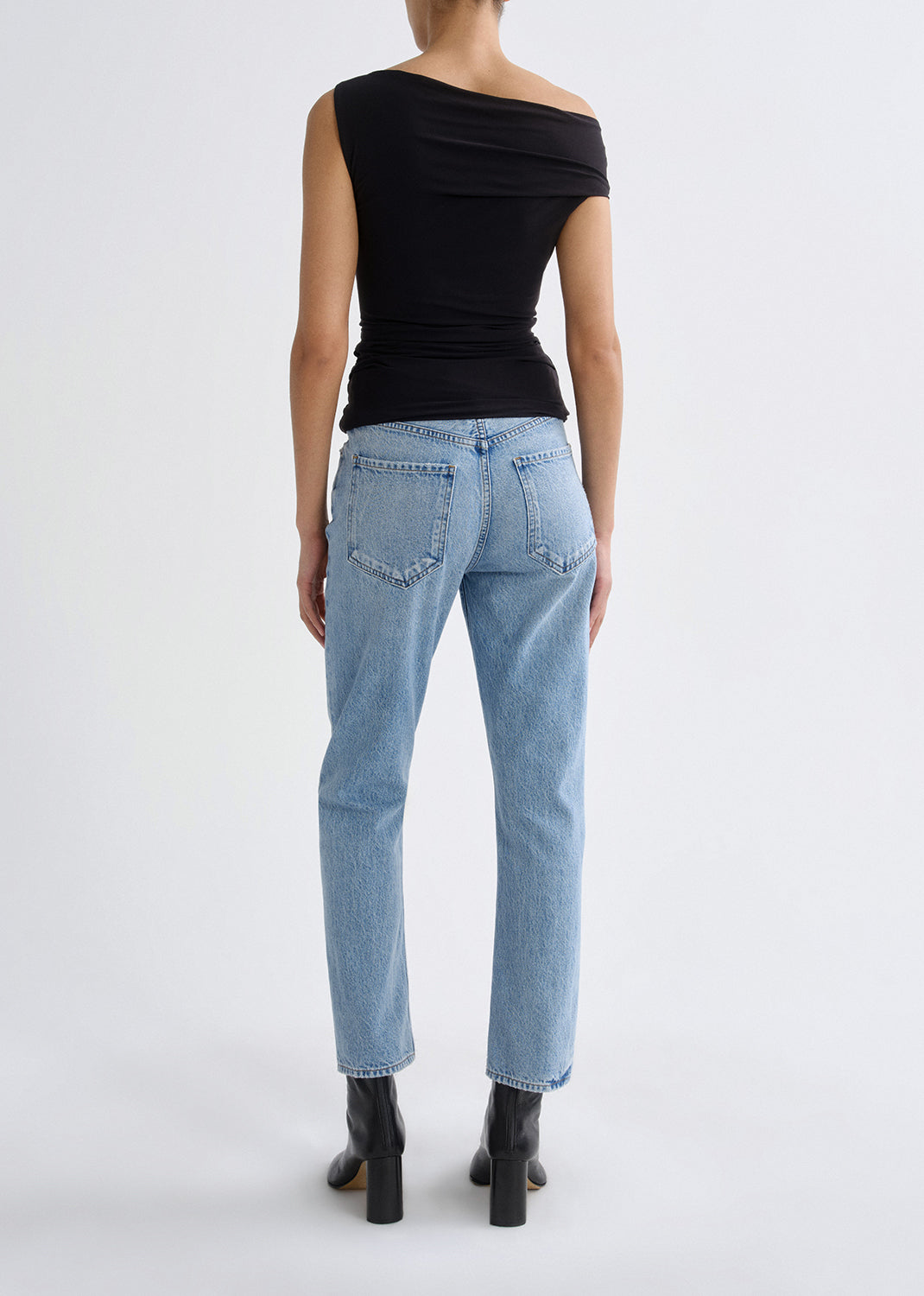 A model shown from the back wearing a black off-the-shoulder top, light-wash blue jeans, and black ankle boots against a plain light gray studio background.