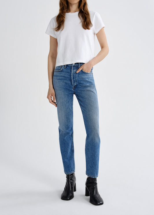 A model wearing a white short-sleeve t-shirt, blue high-waisted straight-leg jeans, and black ankle boots, standing against a light gray studio background.