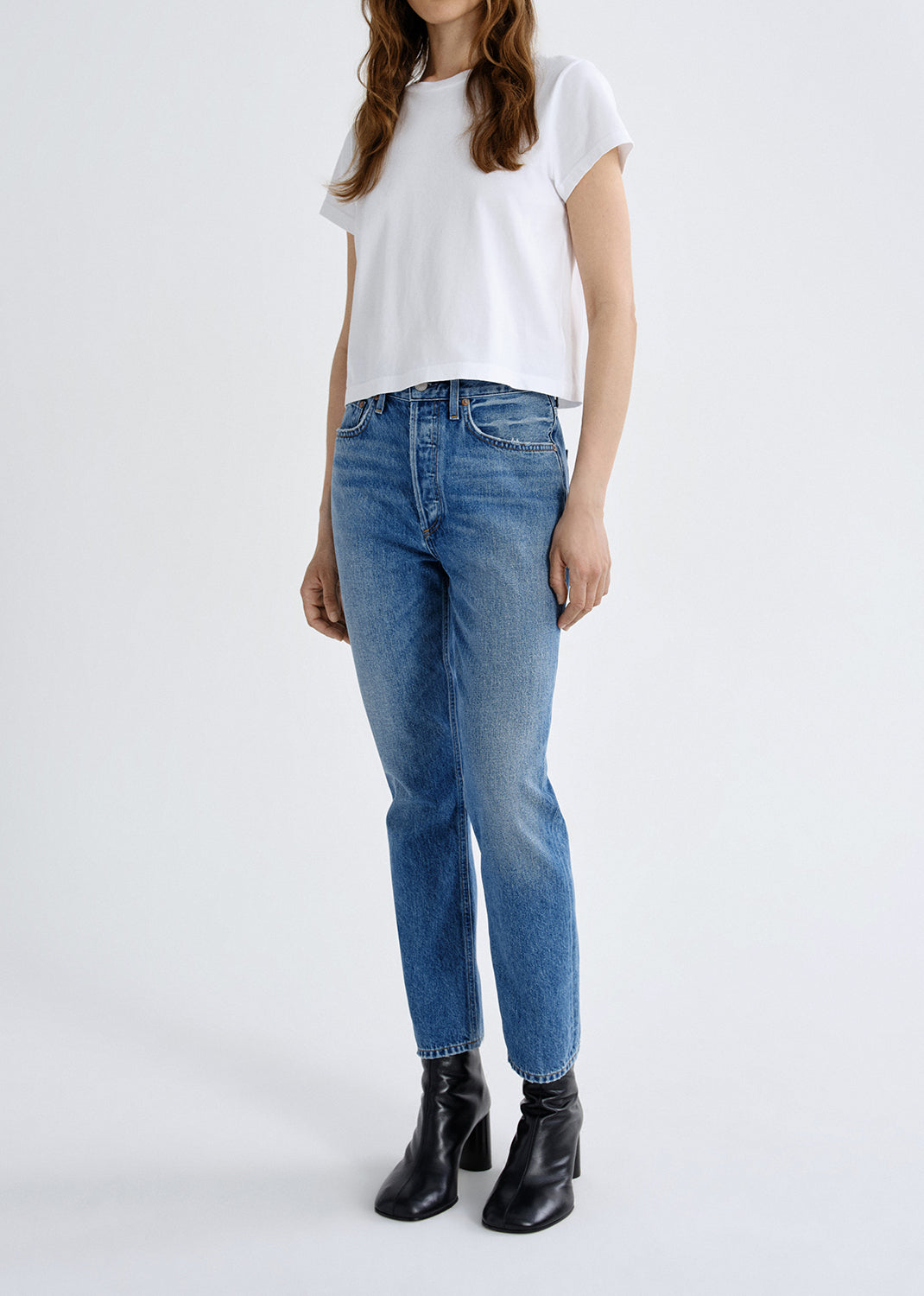 A model wearing a white short-sleeve t-shirt, blue high-waisted straight-leg jeans, and black leather ankle boots, standing against a light gray studio background.