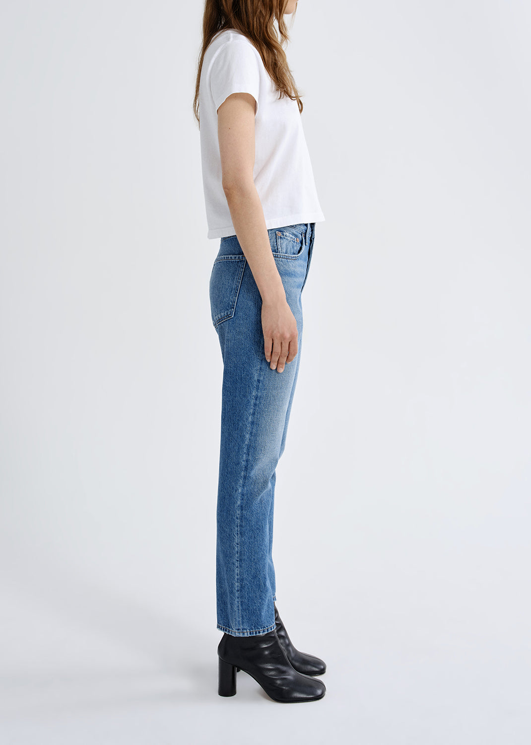 A model shown from the side wearing a white short-sleeve t-shirt, blue straight-leg jeans, and black leather ankle boots against a plain white studio background.
