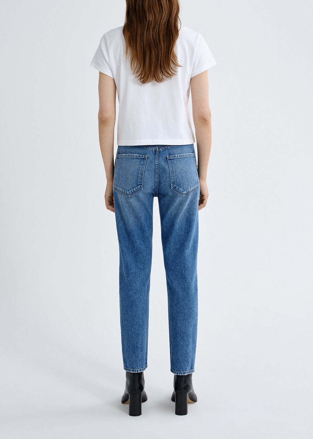 A woman viewed from the back wearing blue straight-leg jeans, a white short-sleeve T-shirt, and black ankle boots against a light gray studio background.