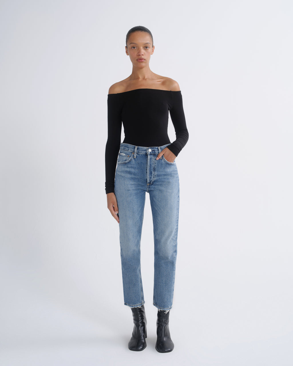 A model stands against a light gray studio background wearing a black off-the-shoulder long-sleeve top, light-wash blue straight-leg jeans, and black ankle boots.