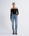 A model stands against a light gray studio background wearing a black off-the-shoulder long-sleeve top, light-wash blue straight-leg jeans, and black ankle boots.
