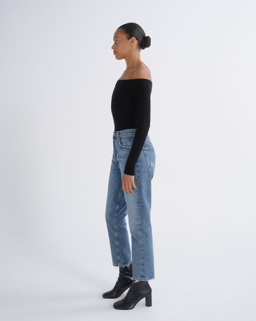 A woman in profile wearing a black off-the-shoulder long-sleeve top, light-wash blue jeans, and black heeled ankle boots, standing against a plain light gray studio background.
