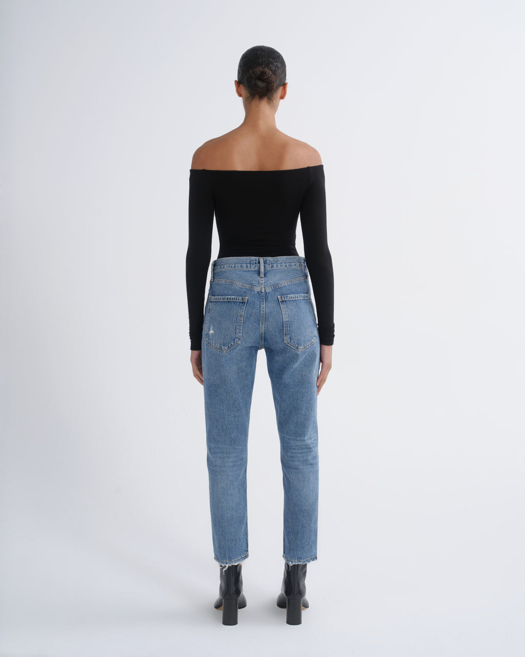 A model viewed from behind wearing a black off-the-shoulder long-sleeve top, light-wash blue jeans, and black ankle boots against a plain light gray studio background.