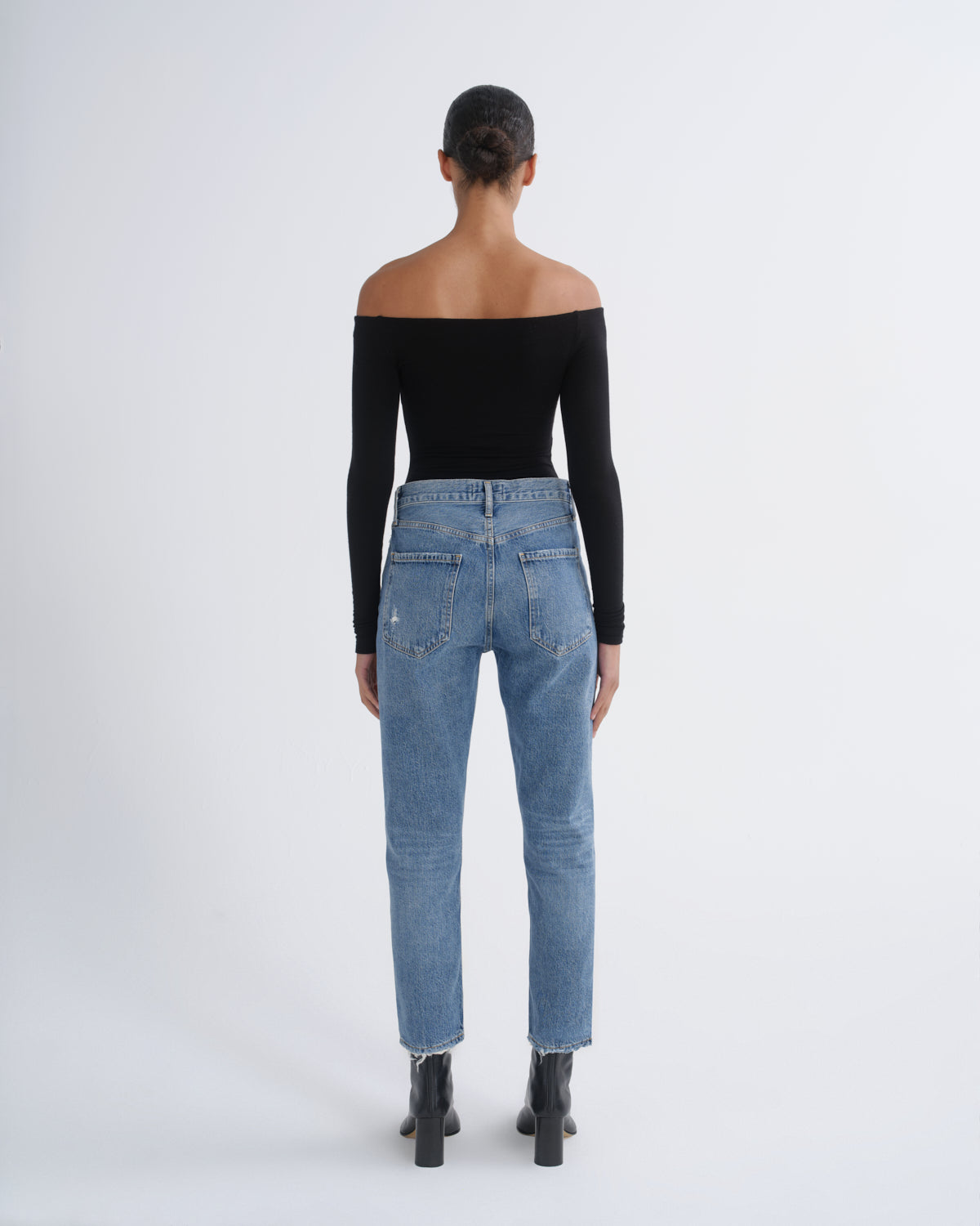 A model viewed from behind wearing a black off-the-shoulder long-sleeve top, light-wash blue jeans, and black ankle boots against a plain light gray studio background.