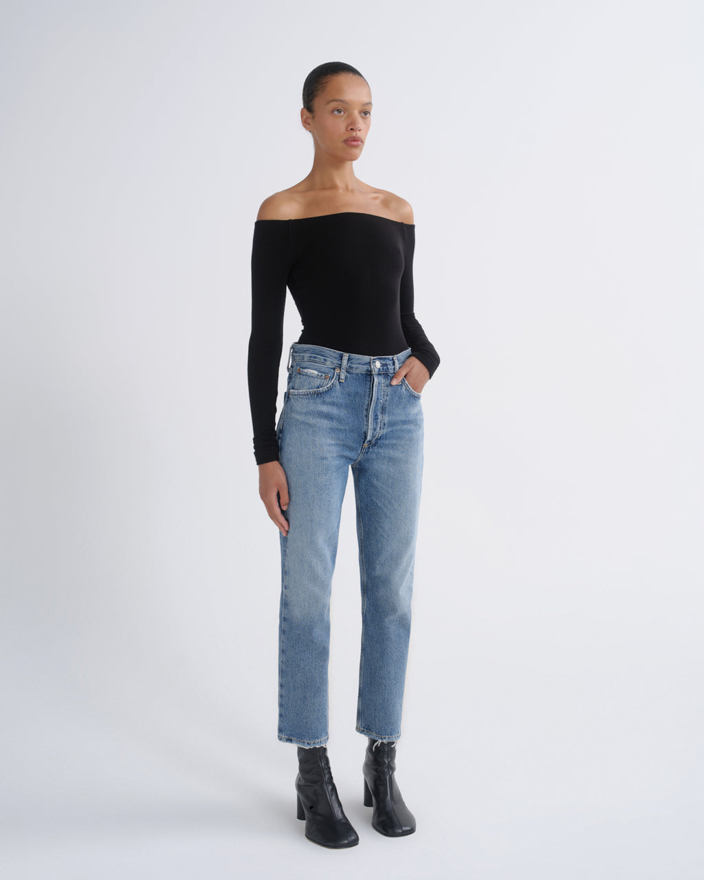 A model stands against a light gray studio background wearing a black off-the-shoulder long-sleeve top, light-wash blue straight-leg jeans, and black ankle boots.
